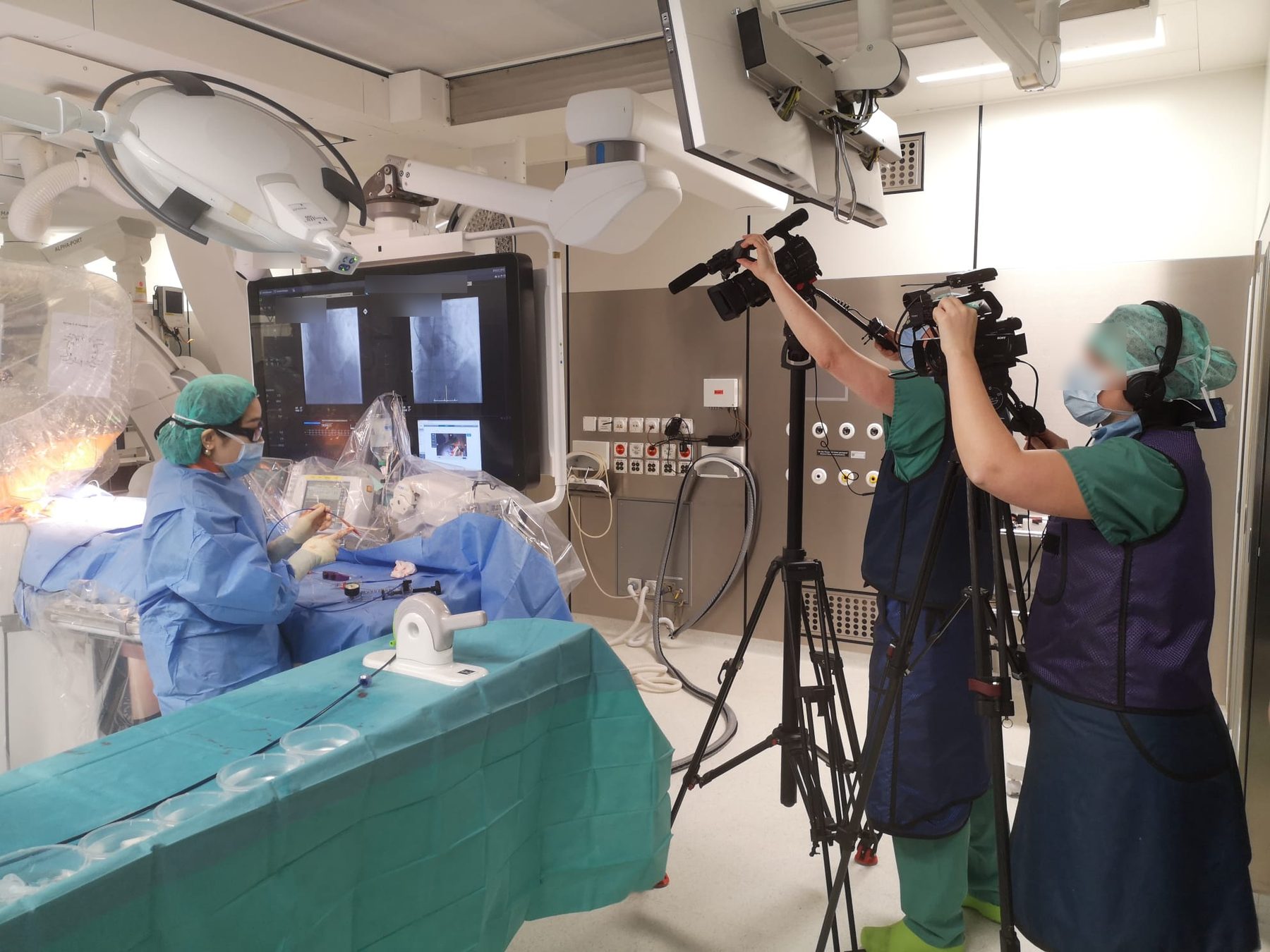 Mi Chen at the table during a live-transmitted structural heart case in Zürich, fluoroscopy screens behind her, broadcast cameras in the foreground